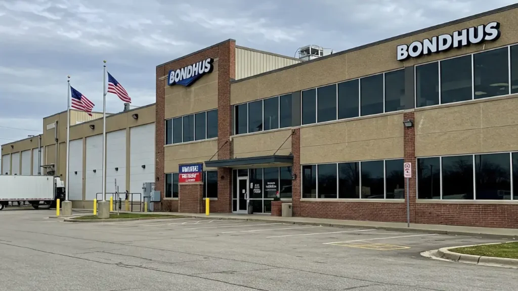 Bondhus manufacturing facility in Monticello, Minnesota, featuring the company's signage on a brick and beige building under a cloudy sky. American flags are visible in front of the building.