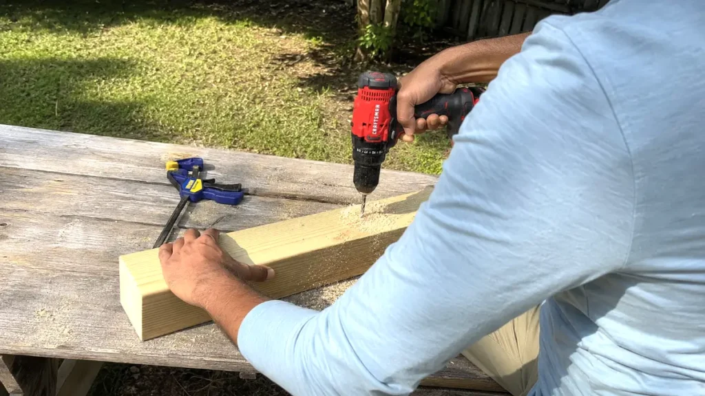 Hands of a person wearing a blue shirt holding a power drill, boring a wide hole into a thick piece of wood.